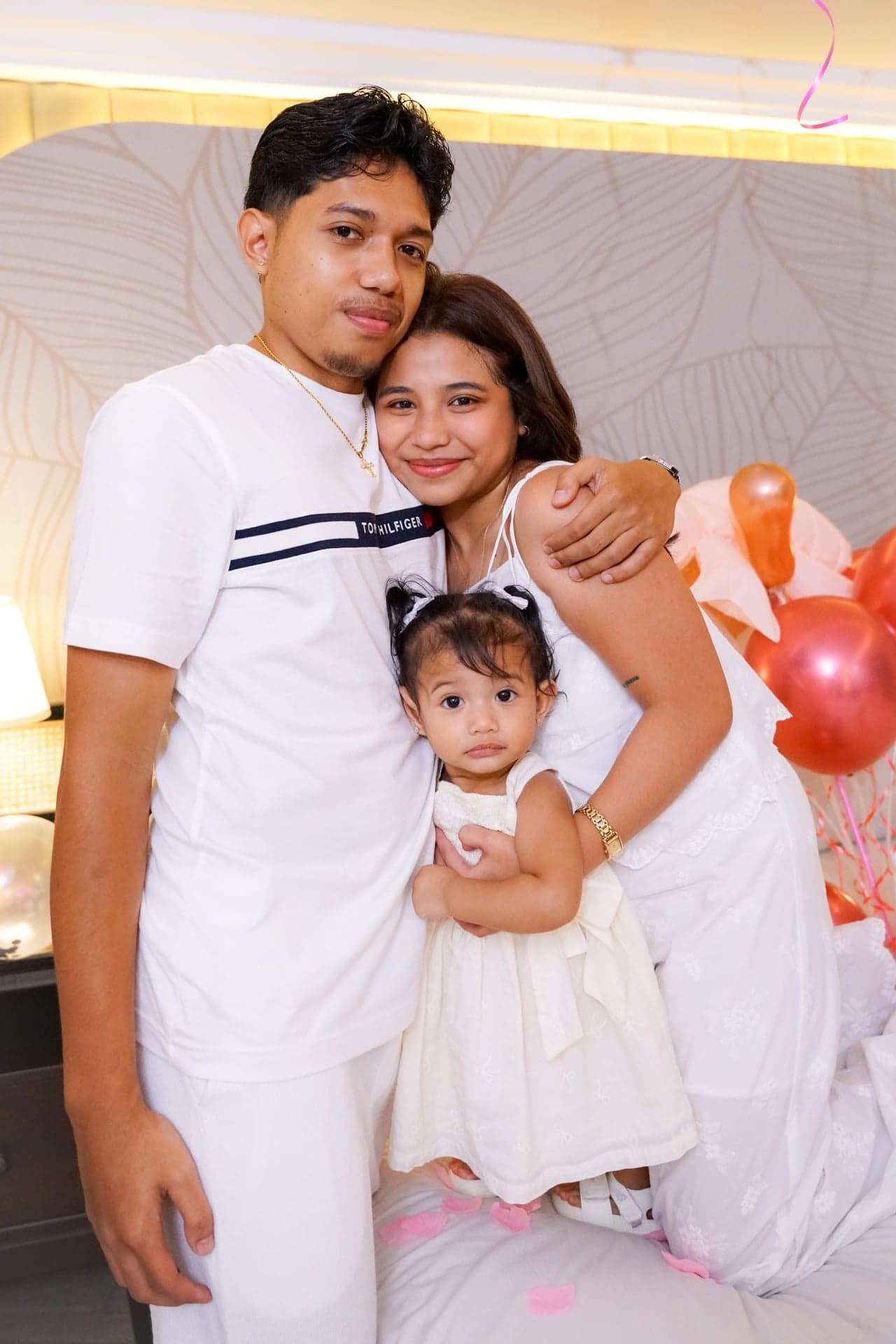 Two adults and a child in white posing together beside balloons