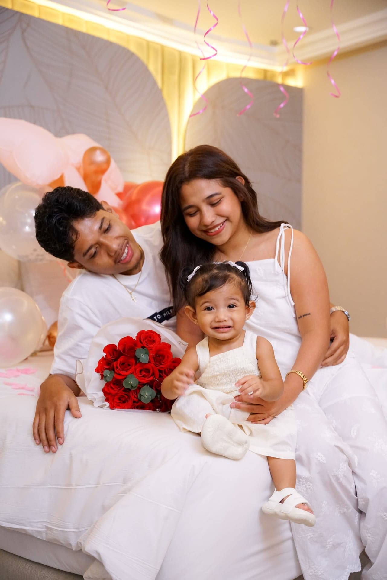 Two adults seated with a child on a bed beside a red rose bouquet