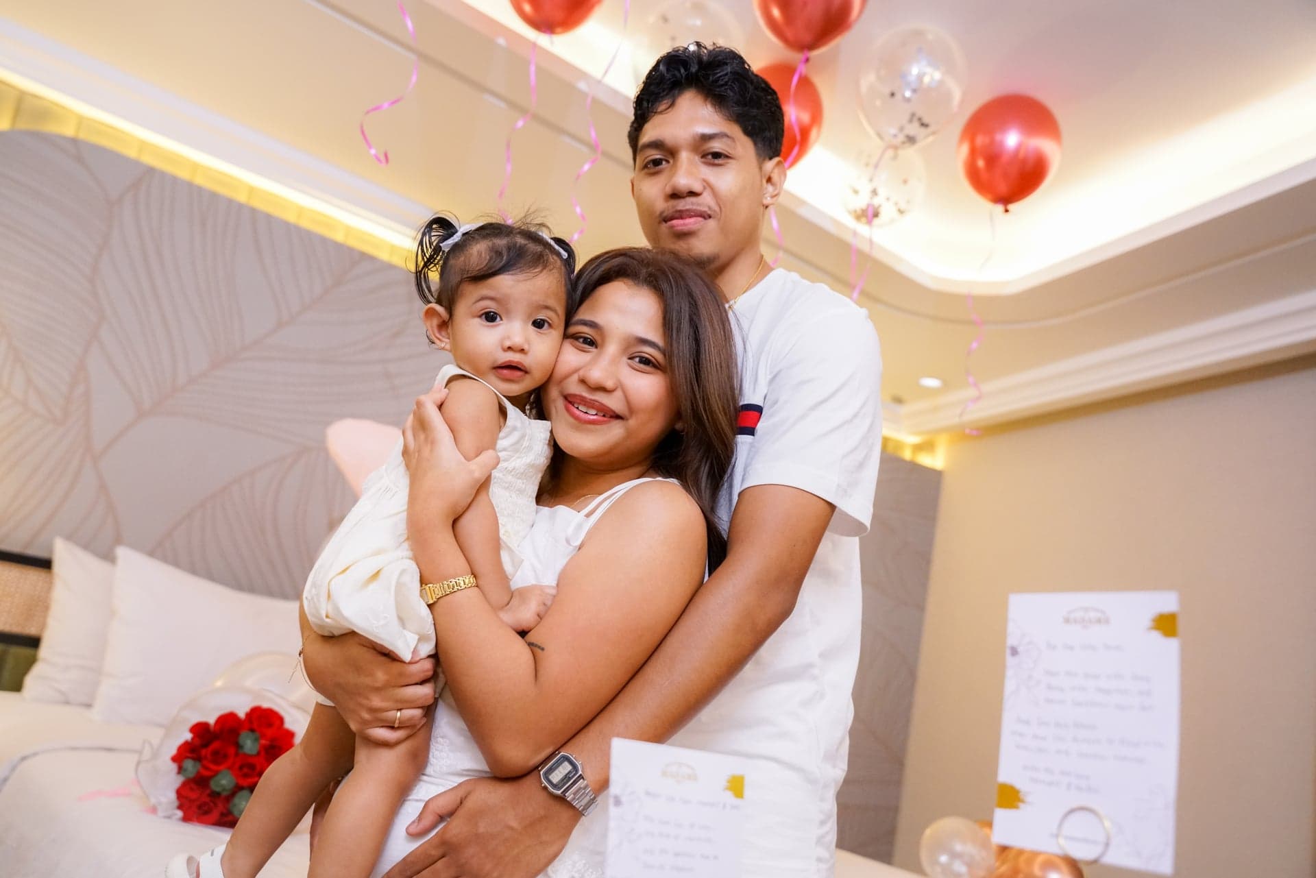 Three people in white embracing with red balloons behind them
