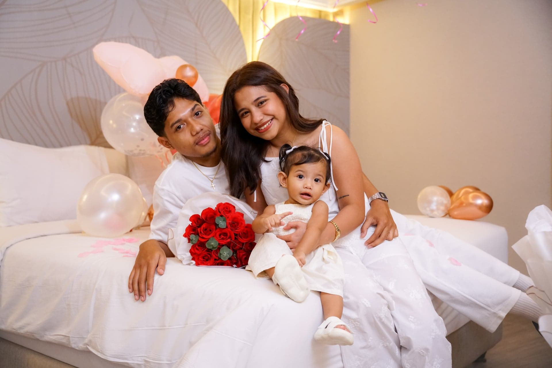 Two adults and a child posing seated on a bed beside red roses